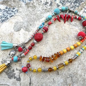 K&ouml;nnte beinhalten: Eine Sammlung farbenfroher Perlenketten mit verschiedenen Anh&auml;ngern und Quasten. Die Halsketten weisen t&uuml;rkisfarbene, rote, gelbe und orangefarbene Perlen sowie silberfarbene Metallelemente auf. Eine Halskette hat einen Buddha-Anh&auml;nger und eine gelbe Quaste.