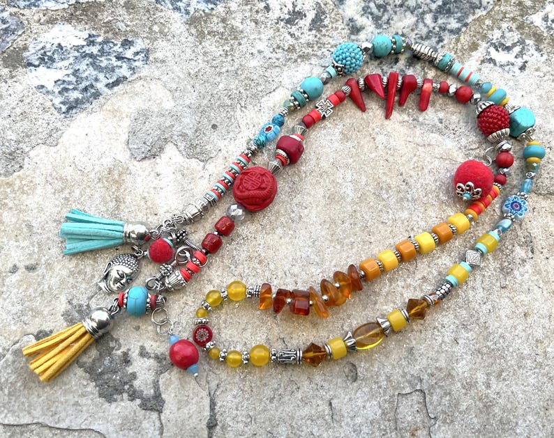 K&ouml;nnte beinhalten: Eine Sammlung farbenfroher Perlenketten mit verschiedenen Anh&auml;ngern und Quasten. Die Halsketten weisen t&uuml;rkisfarbene, rote, gelbe und orangefarbene Perlen sowie silberfarbene Metallelemente auf. Eine Halskette hat einen Buddha-Anh&auml;nger und eine gelbe Quaste.