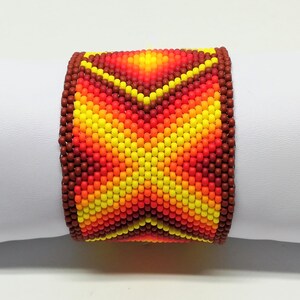 Op de afbeelding: Een brede, met kralen bezette armband met een levendig, geometrisch ontwerp. De armband heeft een kleurengradatie van rood, oranje en geel, met een bruine rand. De kralen zijn klein en strak geweven, waardoor een textuur ontstaat. De armband is ongeveer 5 cm breed.