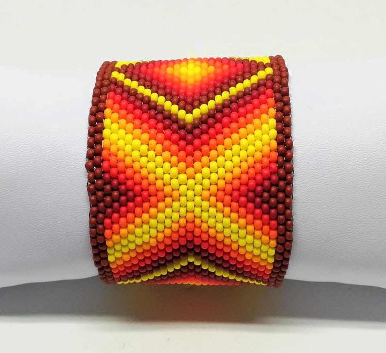 Op de afbeelding: Een brede, met kralen bezette armband met een levendig, geometrisch ontwerp. De armband heeft een kleurengradatie van rood, oranje en geel, met een bruine rand. De kralen zijn klein en strak geweven, waardoor een textuur ontstaat. De armband is ongeveer 5 cm breed.