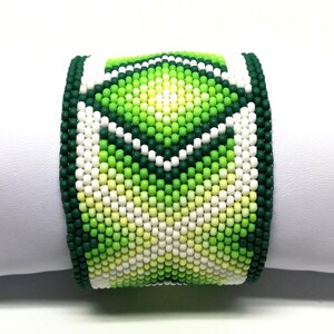 May include: A wide beaded bracelet featuring a geometric pattern in shades of green, white, and yellow. The bracelet is made of small, round beads and has a dark green border. The design is centered on the bracelet.