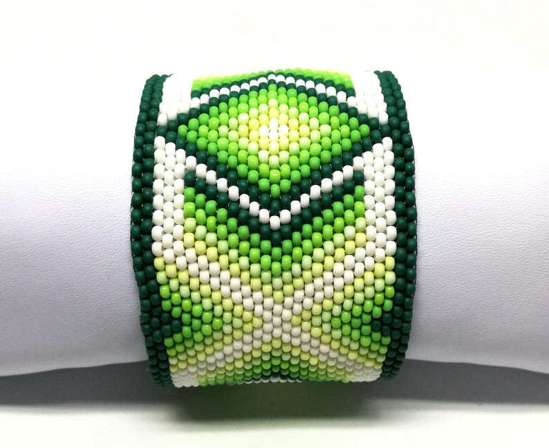 May include: A wide beaded bracelet featuring a geometric pattern in shades of green, white, and yellow. The bracelet is made of small, round beads and has a dark green border. The design is centered on the bracelet.