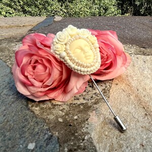 May include: A white beaded brooch with a sculpted face design, pinned to a pink silk rose.