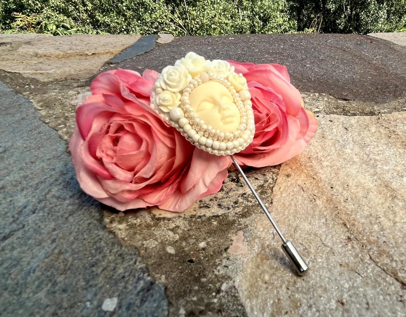 May include: A white beaded brooch with a sculpted face design, pinned to a pink silk rose.