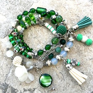 May include: A collection of green beaded bracelets with various charms and tassels. The bracelets feature a mix of glass, stone, and metal beads in shades of green, white, and silver. Some bracelets have decorative charms and tassels.