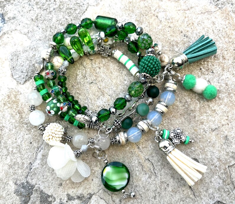 May include: A collection of green beaded bracelets with various charms and tassels. The bracelets feature a mix of glass, stone, and metal beads in shades of green, white, and silver. Some bracelets have decorative charms and tassels.