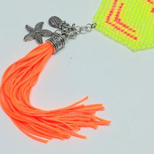 May include: Vibrant orange tassel with silver charms, including a starfish and pineapple. The tassel is attached to a small, beaded, neon yellow and pink object. The charms and tassel are on a white background.