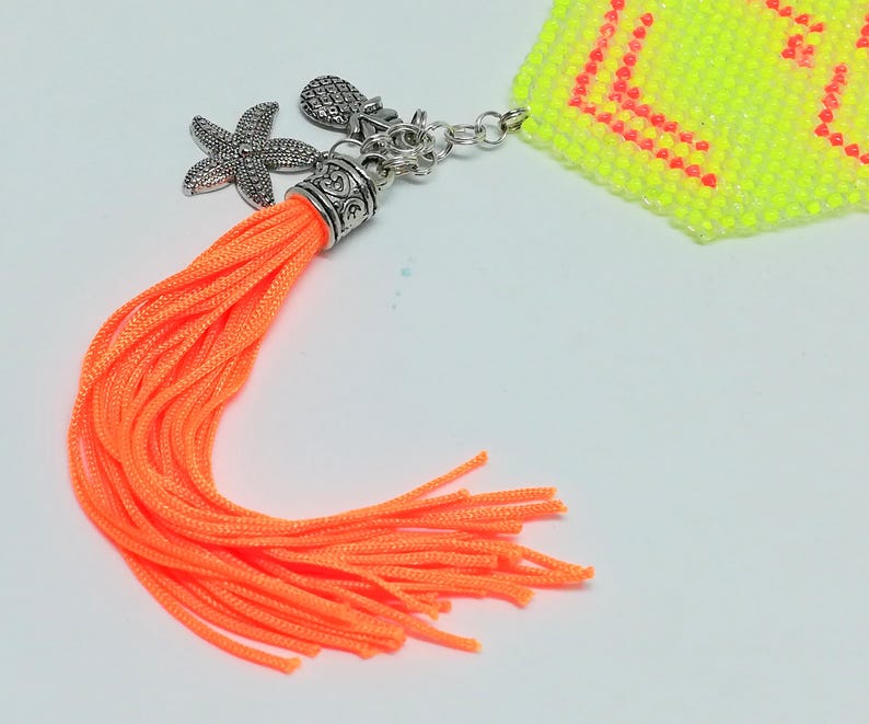 May include: Vibrant orange tassel with silver charms, including a starfish and pineapple. The tassel is attached to a small, beaded, neon yellow and pink object. The charms and tassel are on a white background.