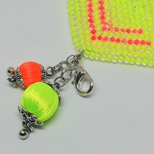 May include: A close-up of a beaded charm with a lobster clasp. The charm features two silk thread tassels in neon orange and green, with silver-coloured metal accents. The background includes a neon yellow beaded section with the letter "L" in red.