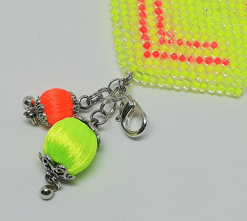 May include: A close-up of a beaded charm with a lobster clasp. The charm features two silk thread tassels in neon orange and green, with silver-coloured metal accents. The background includes a neon yellow beaded section with the letter "L" in red.