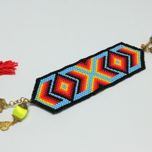 May include: A beaded bookmark with a geometric design in red, orange, yellow, and blue, outlined in black. It has a red tassel on one end and decorative charms on the other, including a yellow bead with a gold wing charm.