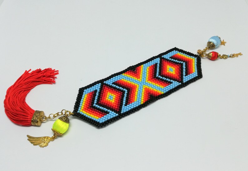 May include: A beaded bookmark with a geometric design in red, orange, yellow, and blue, outlined in black. It has a red tassel on one end and decorative charms on the other, including a yellow bead with a gold wing charm.