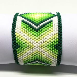 May include: A wide beaded bracelet featuring a geometric design in shades of green, white, and yellow. The bracelet is primarily green with a dark green border and a central pattern of lighter green and white beads. The bracelet is displayed on a white surface.