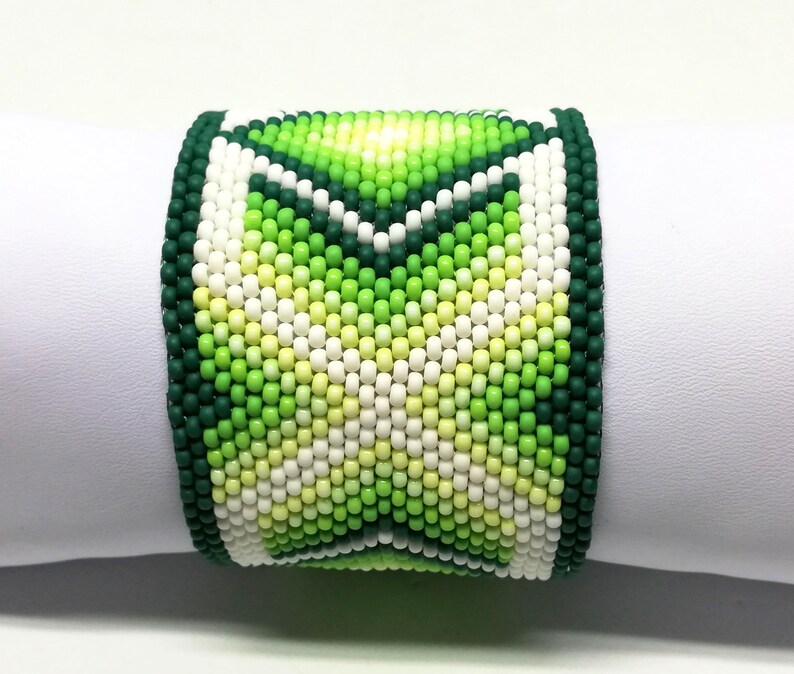 May include: A wide beaded bracelet featuring a geometric design in shades of green, white, and yellow. The bracelet is primarily green with a dark green border and a central pattern of lighter green and white beads. The bracelet is displayed on a white surface.