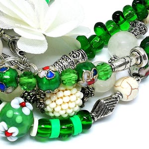 May include: Close-up of beaded bracelets featuring green, white, and silver accents. The bracelets include various bead shapes and sizes, with floral and geometric designs. A white flower is visible in the background.
