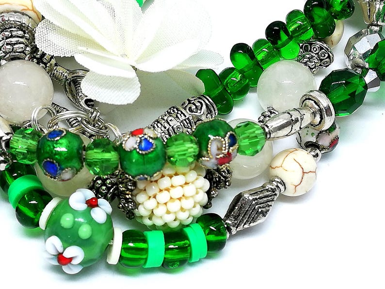 May include: Close-up of beaded bracelets featuring green, white, and silver accents. The bracelets include various bead shapes and sizes, with floral and geometric designs. A white flower is visible in the background.