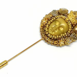May include: A gold-toned stick pin with a detailed, circular design. The pin features a central face, surrounded by beads, small flowers, and leaves. A small crown and decorative beads accent the design. The pin is set against a white background.
