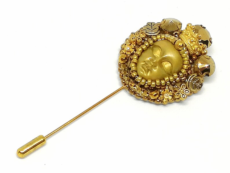 May include: A gold-toned stick pin with a detailed, circular design. The pin features a central face, surrounded by beads, small flowers, and leaves. A small crown and decorative beads accent the design. The pin is set against a white background.