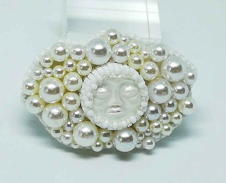May include: A white pearl brooch with a white face in the center. The face has closed eyes and a small mouth.