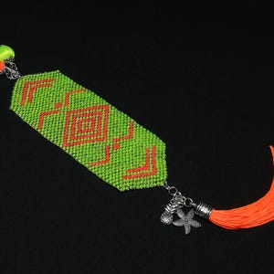 May include: A beaded bracelet with a lime green base and a geometric orange pattern. The bracelet has a silver chain with charms and an orange tassel. The bracelet is approximately 3.8 cm wide.
