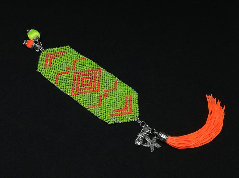 May include: A beaded bracelet with a lime green base and a geometric orange pattern. The bracelet has a silver chain with charms and an orange tassel. The bracelet is approximately 3.8 cm wide.