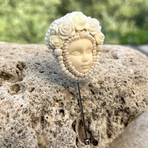 May include: A white porcelain pin with a face and a crown of white roses. The pin is beaded with white pearls and is attached to a silver pin.