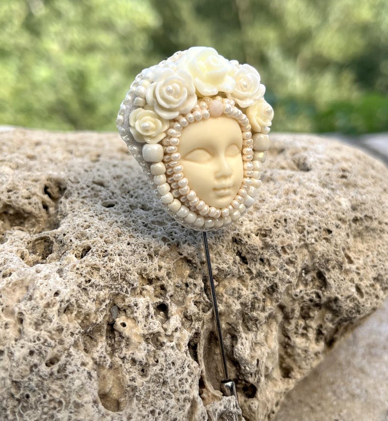 May include: A white porcelain pin with a face and a crown of white roses. The pin is beaded with white pearls and is attached to a silver pin.