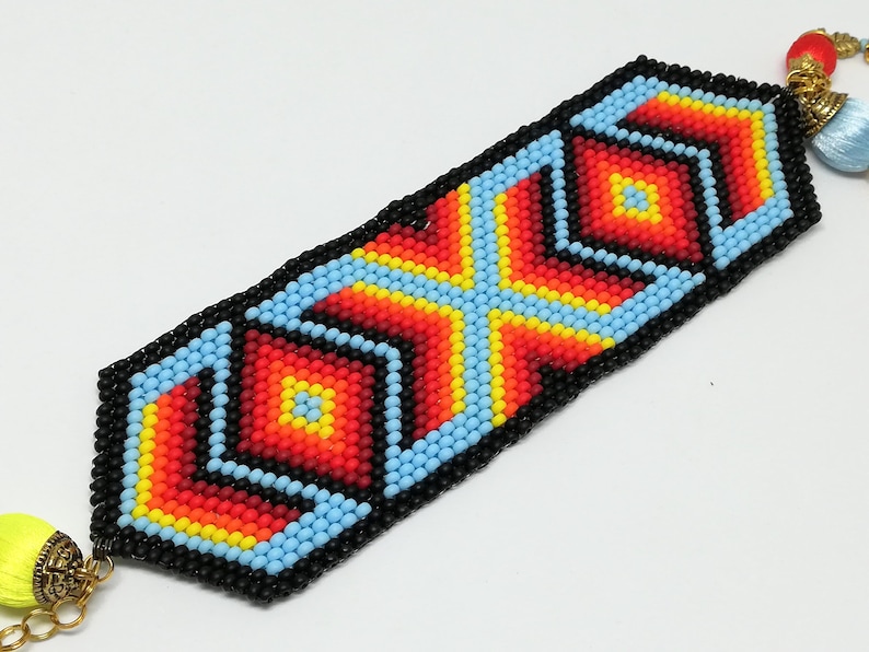 May include: A beaded bracelet with a geometric design. The bracelet features a black border with a central pattern of red, orange, yellow, and blue beads. Tassels and gold-colored chain are attached to the ends.