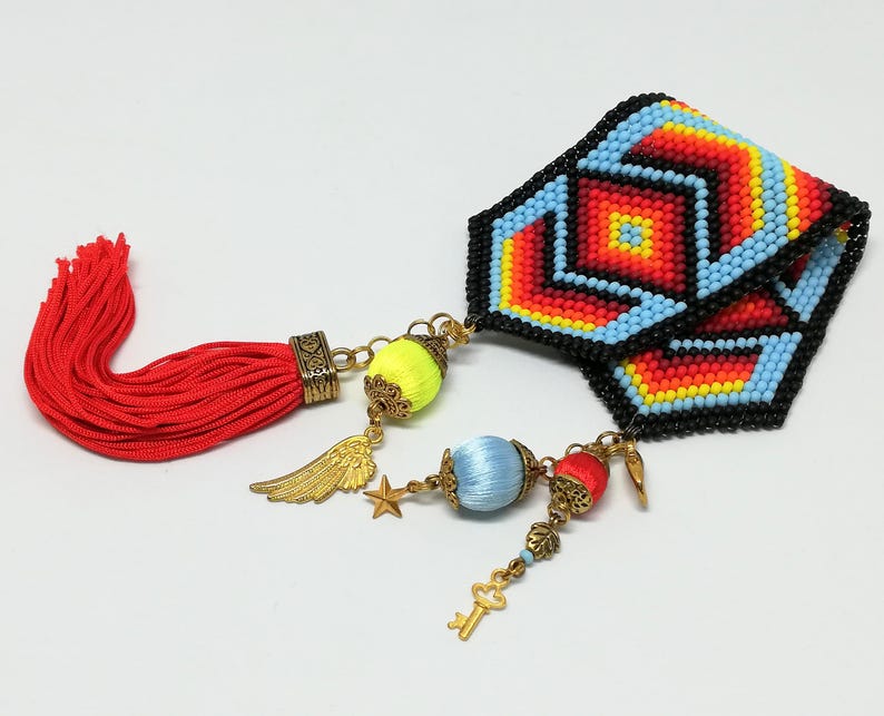 May include: A beaded bracelet with a geometric pattern in red, orange, yellow, and blue. It features a red tassel and decorative charms, including a wing, star, and key, with yellow and blue accents.