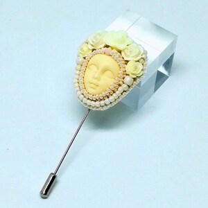 May include: A delicate brooch featuring a sculpted face surrounded by white and yellow flowers and pearls. The brooch is attached to a silver pin.