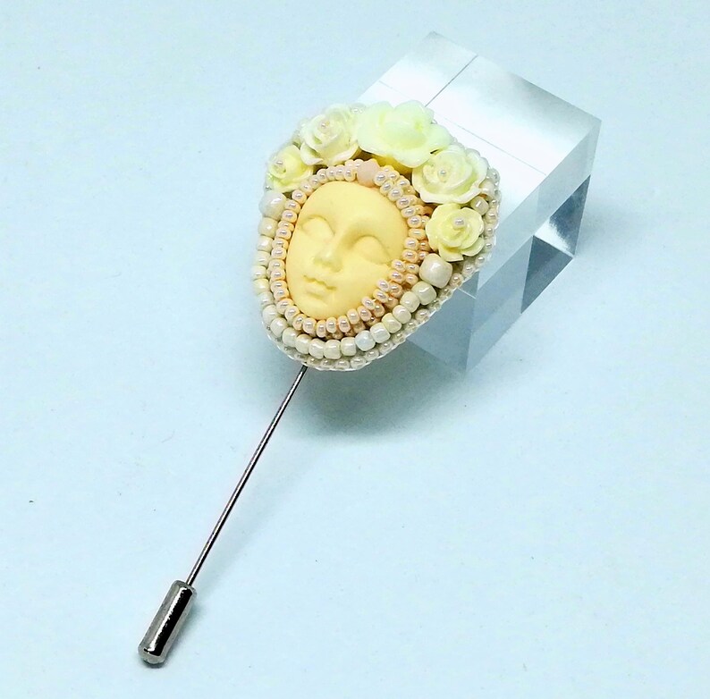 May include: A delicate brooch featuring a sculpted face surrounded by white and yellow flowers and pearls. The brooch is attached to a silver pin.