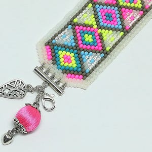 May include: A beaded bracelet with a geometric pattern in pink, yellow, blue, and gray. The bracelet has a silver clasp and a pink silk charm with a silver heart and leaf-shaped pendant. The bracelet is approximately 1 inch wide.