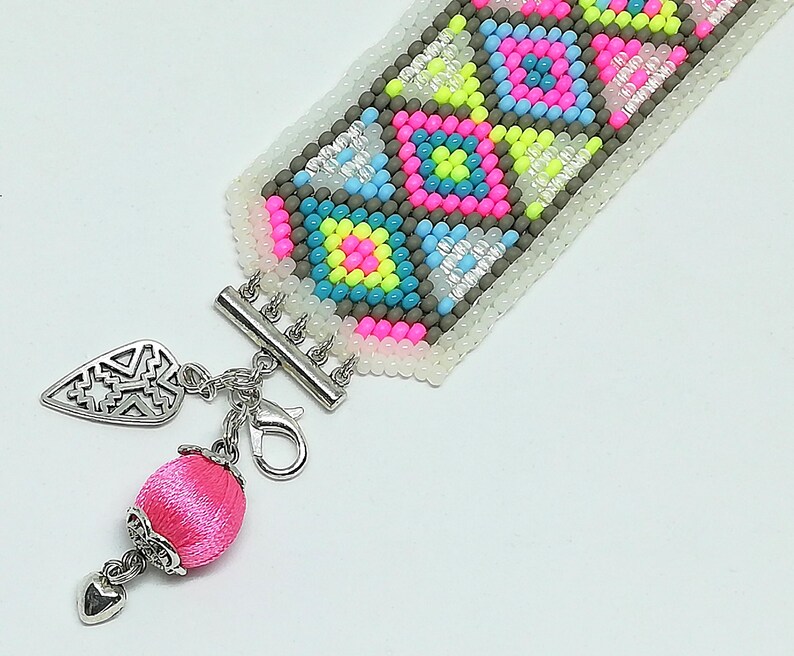 May include: A beaded bracelet with a geometric pattern in pink, yellow, blue, and gray. The bracelet has a silver clasp and a pink silk charm with a silver heart and leaf-shaped pendant. The bracelet is approximately 1 inch wide.
