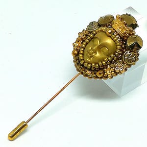 May include: A gold-toned stick pin featuring a detailed face with a crown, surrounded by beads and decorative elements. The pin has a long, slender shaft and a rounded end. The design incorporates leaves and floral accents, creating an ornate and regal appearance.