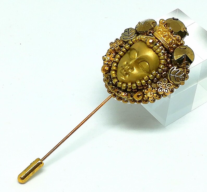 May include: A gold-toned stick pin featuring a detailed face with a crown, surrounded by beads and decorative elements. The pin has a long, slender shaft and a rounded end. The design incorporates leaves and floral accents, creating an ornate and regal appearance.