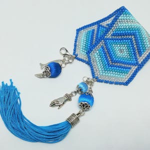 May include: A handmade blue and silver beaded bookmark with a geometric design. The bookmark features a tassel and decorative charms, including a hand and a bell-shaped charm. The color palette includes shades of blue and silver.