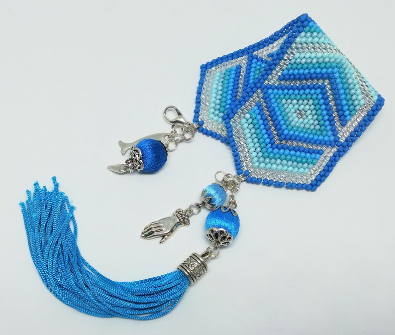 May include: A handmade blue and silver beaded bookmark with a geometric design. The bookmark features a tassel and decorative charms, including a hand and a bell-shaped charm. The color palette includes shades of blue and silver.