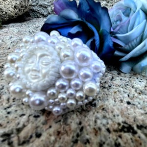 May include: A white sun face with closed eyes surrounded by white pearls, a decorative piece.