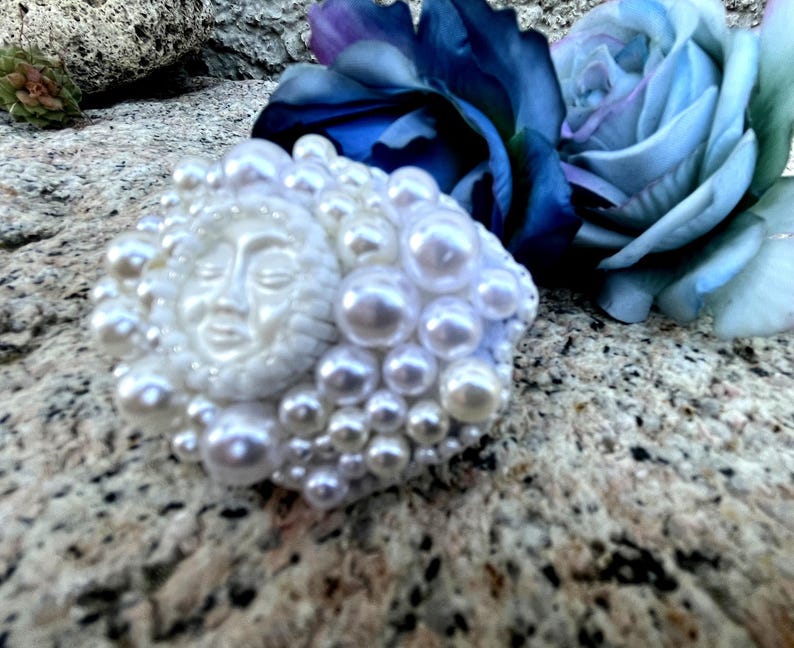 May include: A white sun face with closed eyes surrounded by white pearls, a decorative piece.