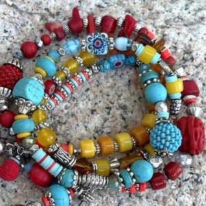 K&ouml;nnte beinhalten: Eine Sammlung farbenfroher Armb&auml;nder mit Perlen. Die Armb&auml;nder weisen eine Vielzahl von Perlen in T&uuml;rkis, Rot, Gelb und Blau mit silbernen Akzenten auf. Einige Perlen haben komplizierte Muster, die einen Bohemian-Stil erzeugen.