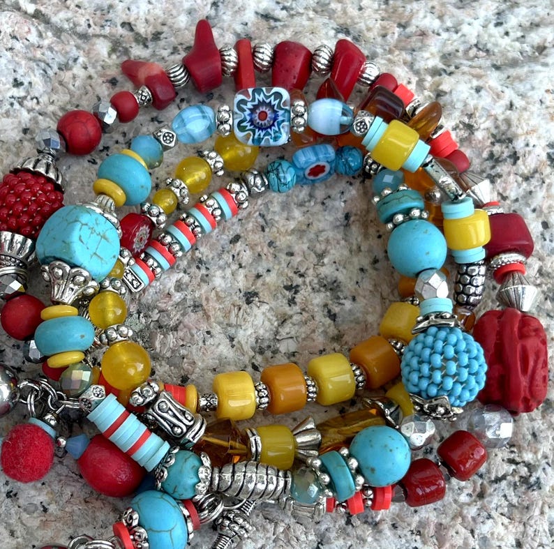 K&ouml;nnte beinhalten: Eine Sammlung farbenfroher Armb&auml;nder mit Perlen. Die Armb&auml;nder weisen eine Vielzahl von Perlen in T&uuml;rkis, Rot, Gelb und Blau mit silbernen Akzenten auf. Einige Perlen haben komplizierte Muster, die einen Bohemian-Stil erzeugen.