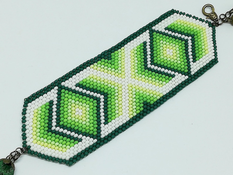 May include: A beaded bracelet featuring a geometric design in shades of green, white, and yellow. The bracelet is rectangular with a dark green border and a bronze-colored clasp. The pattern includes diamond and cross shapes.