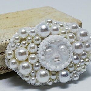 May include: A white pearl brooch with a sculpted face in the center. The face has closed eyes and a serene expression. The brooch is surrounded by a cluster of white pearls.