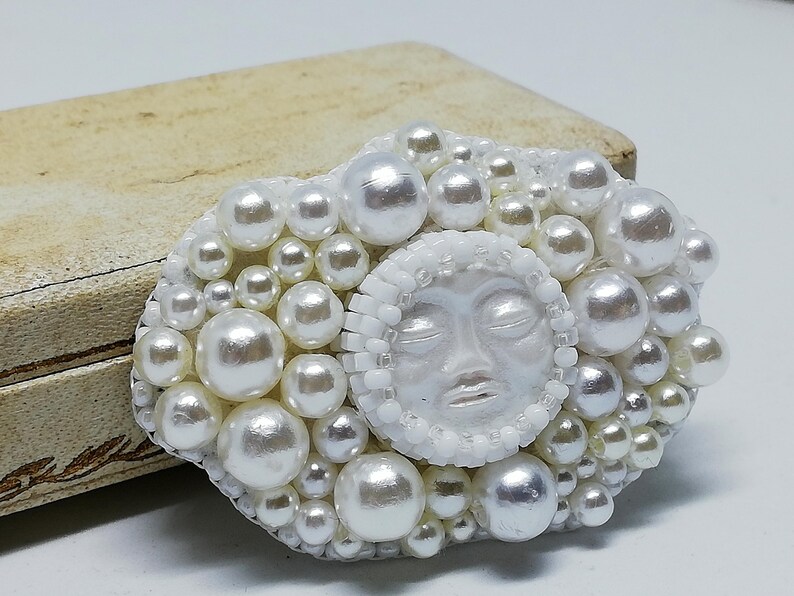 May include: A white pearl brooch with a sculpted face in the center. The face has closed eyes and a serene expression. The brooch is surrounded by a cluster of white pearls.