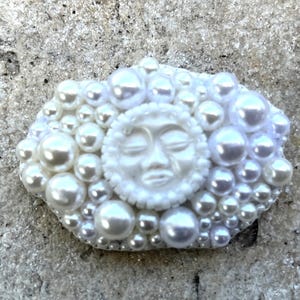 May include: A white ceramic sun with a face sculpted on it, surrounded by white pearls.