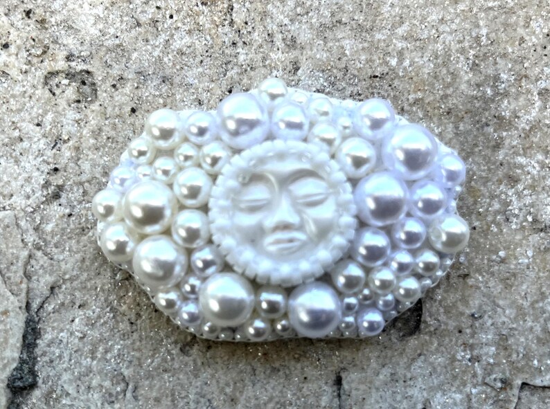 May include: A white ceramic sun with a face sculpted on it, surrounded by white pearls.
