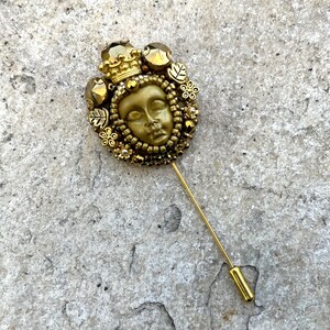 May include: A gold-toned stick pin featuring a detailed face design, surrounded by a crown, leaves, and floral accents. The pin has a long, slender shaft and a simple, cylindrical end. The overall design is ornate and vintage-inspired.
