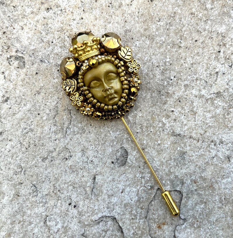 May include: A gold-toned stick pin featuring a detailed face design, surrounded by a crown, leaves, and floral accents. The pin has a long, slender shaft and a simple, cylindrical end. The overall design is ornate and vintage-inspired.