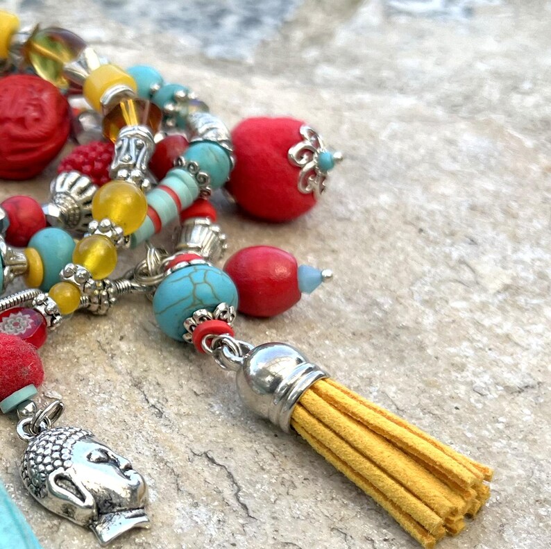K&ouml;nnte beinhalten: Nahaufnahme einer Perlenkette mit einem silbernen Buddha-Anh&auml;nger und einer gelben Quaste. Die Halskette hat rote, t&uuml;rkisfarbene und gelbe Perlen. Die Perlen sind mit silbernen Metallakzenten aufgereiht. Der Hintergrund ist eine hellgraue Steinoberfl&auml;che.