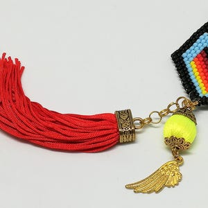 May include: Close-up of a beaded pendant with a red tassel. The pendant features a geometric design in black, blue, red, and yellow. A gold-colored wing charm and a neon yellow ball are attached.